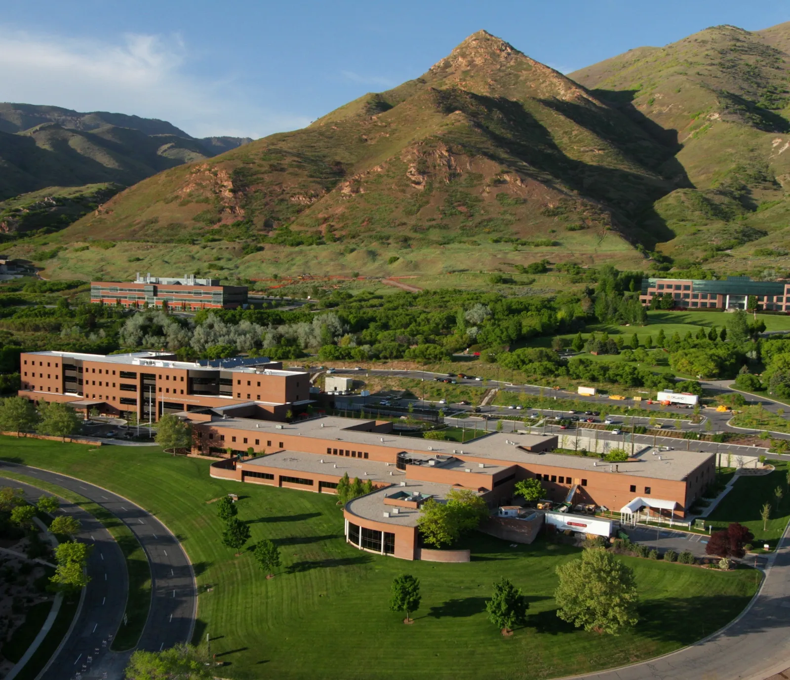 Aerial view of a modern campus with brick buildings, green lawns, and trees, set against a backdrop of large, rugged mountains under a clear blue sky.