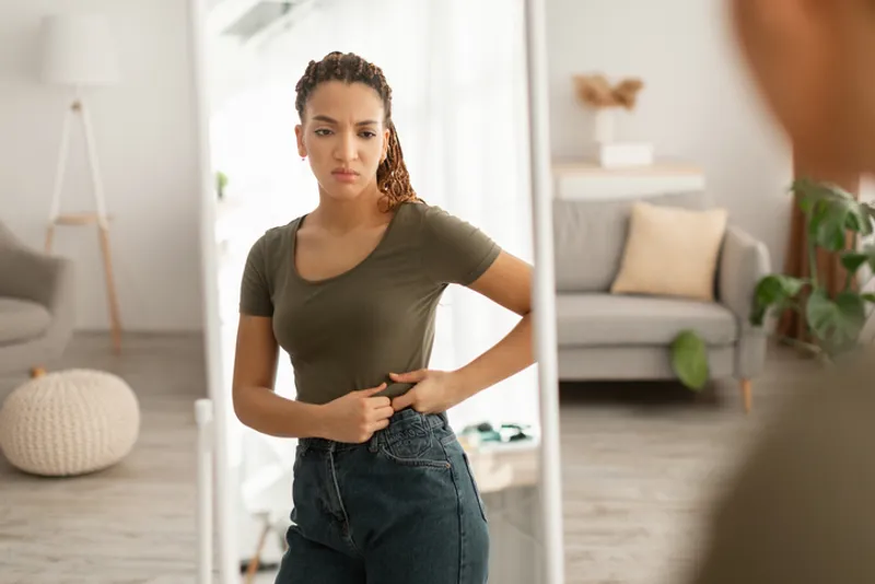 Unsuccessful Slimming. Discontented African American Lady Touching Fat Belly After Weight Gain Looking At Mirror Standing At Home. Excess Weight Problem Concept. Selective Focus