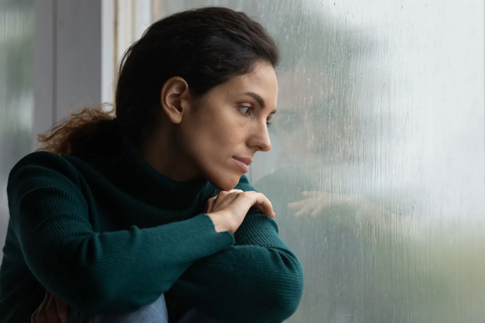Thoughtful stressed young hispanic latin woman sitting on windowsill, looking outside on rainy weather, having depressive or melancholic mood, suffering from negative thoughts alone at home.