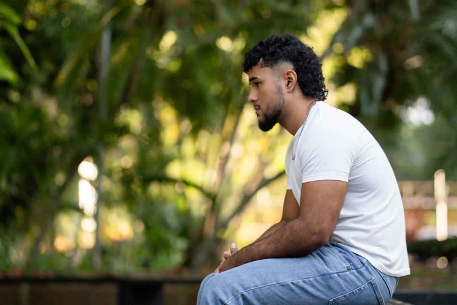 Young man sitting in profile in a park setting, contemplating with a thoughtful expression. Concept of solitude and reflection