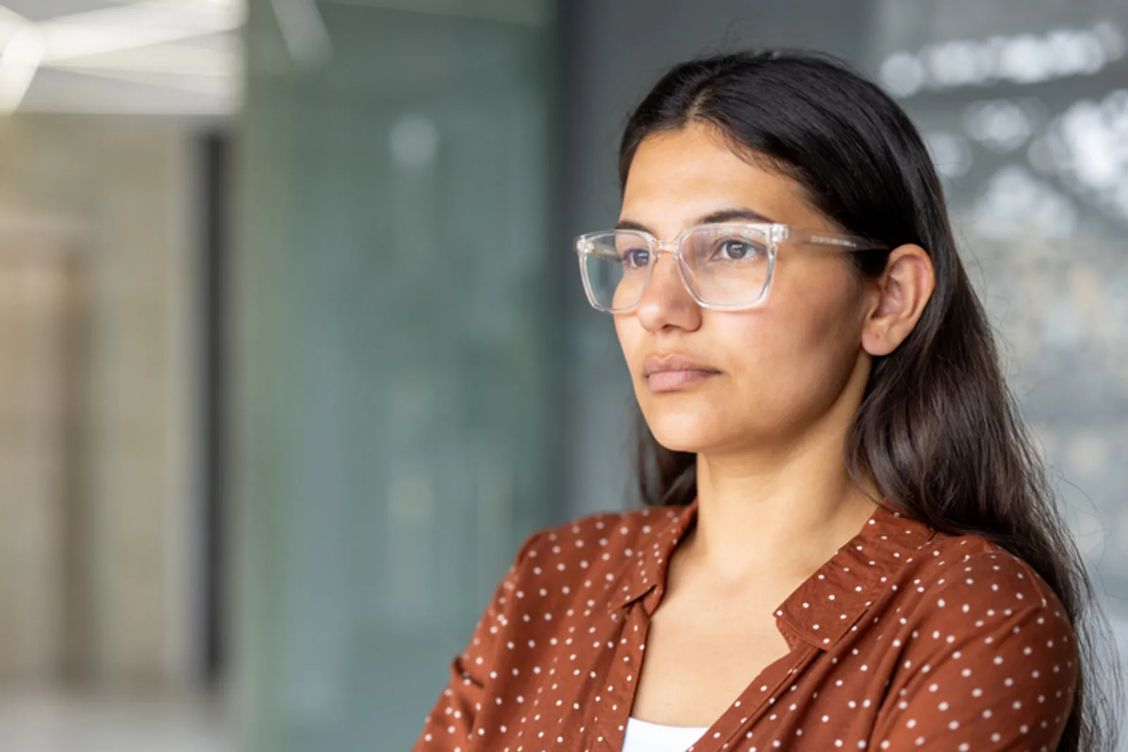 Confident indian woman wearing clear frame glasses looks away, showing a thoughtful and determined expression in a corporate office setting, representing career growth and empowerment