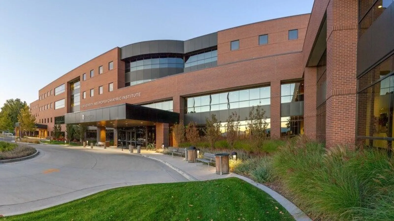 A modern brick building with large glass windows, the sign reads University Neuropsychiatric Institute. There is a curved driveway, green grass, shrubs, and trees in front, with clear blue skies above.
