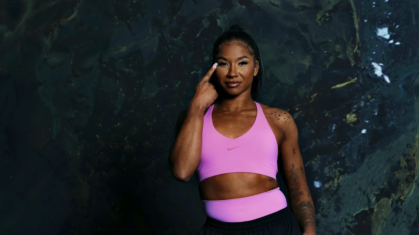 A woman in a pink sports bra and black pants stands against a dark marbled background, pointing a to her head.