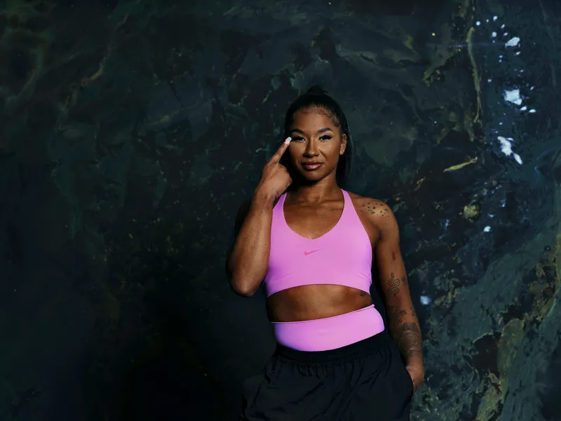 A woman in a pink sports bra and black pants stands against a dark marbled background, making a heart shape with her hands.