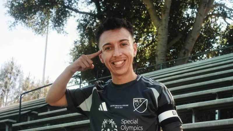 A smiling soccer player in uniform sits on bleachers, resting one foot on a soccer ball. He points to his temple and wears a captains armband. Trees and a field are visible in the background.