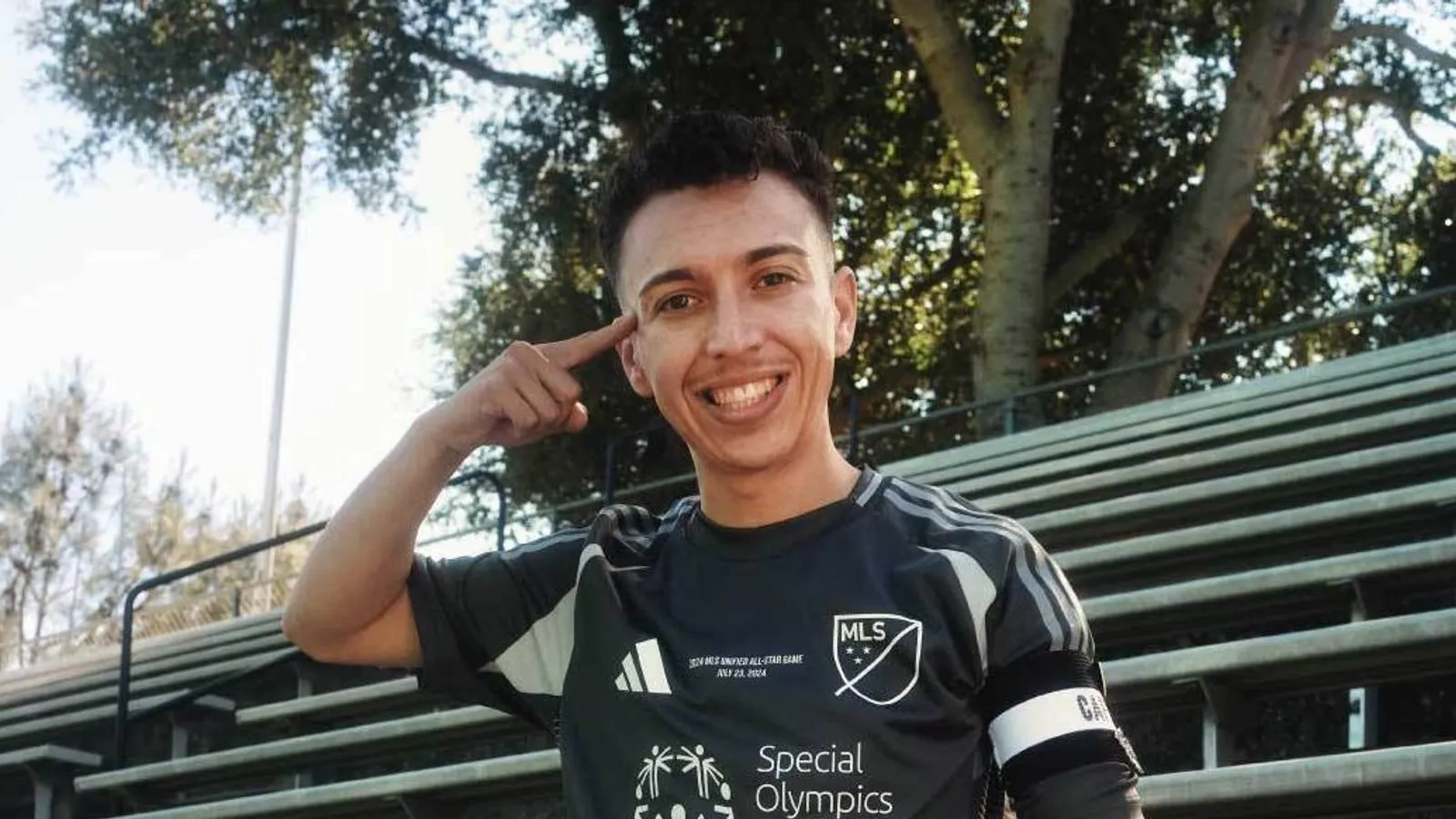 A smiling soccer player in uniform sits on bleachers, resting one foot on a soccer ball. He points to his temple and wears a captains armband. Trees and a field are visible in the background.
