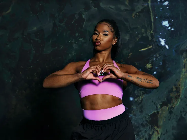 A woman in a pink sports bra and black pants stands against a dark marbled background, making a heart shape with her hands.